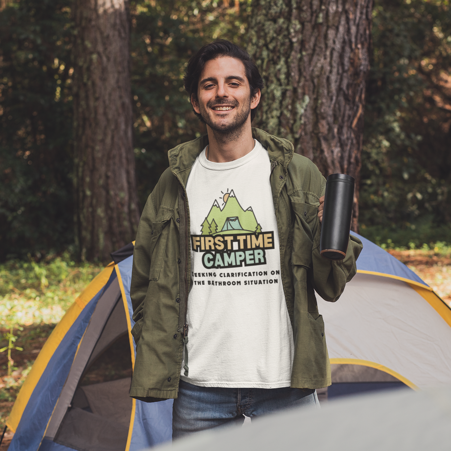 Man wearing a 'First-Time Camper' t-shirt standing next to a tent in a forest.