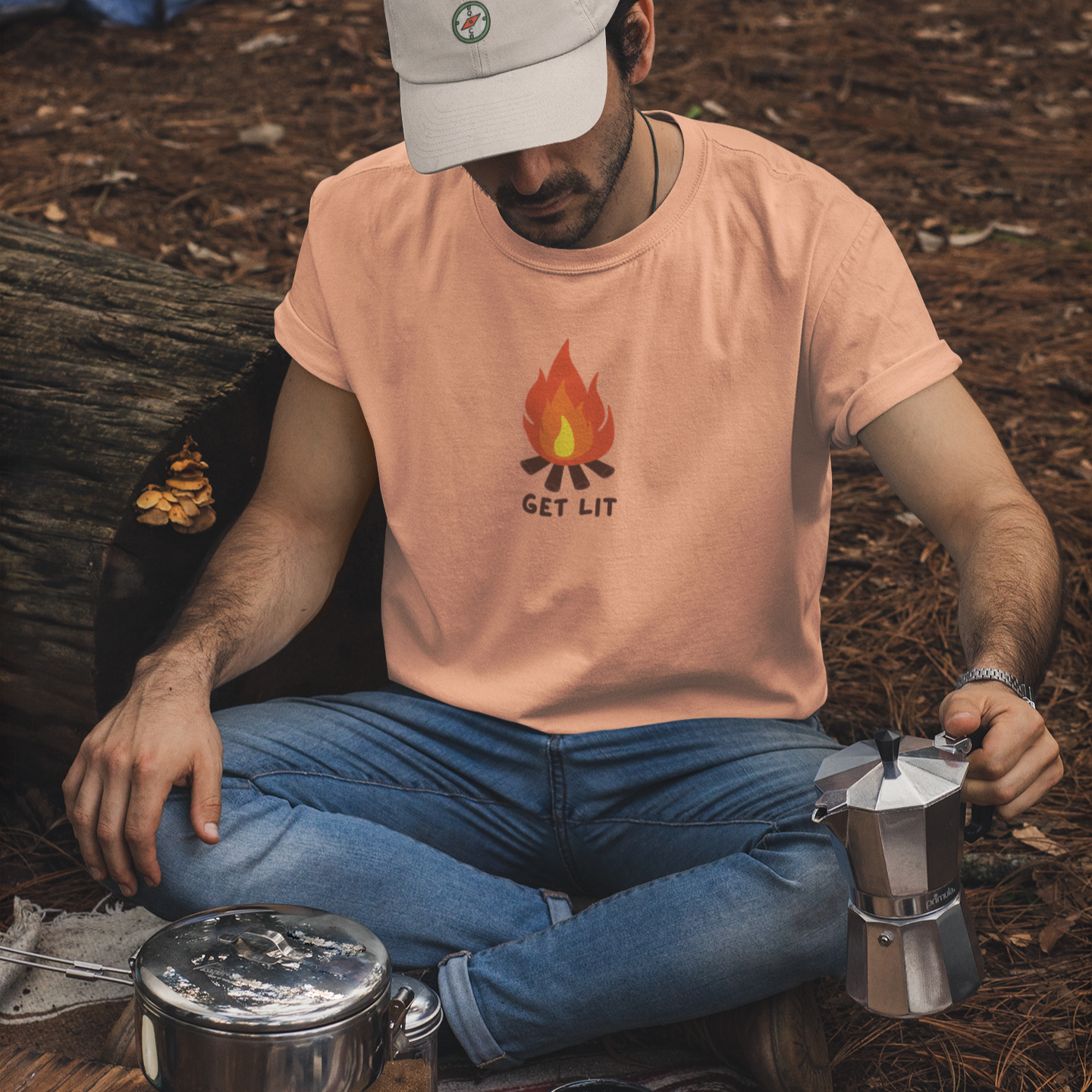 Man camping, sitting by a campfire with a Moka pot and coffee supplies wearing an outdoor shirt that says Get Lit