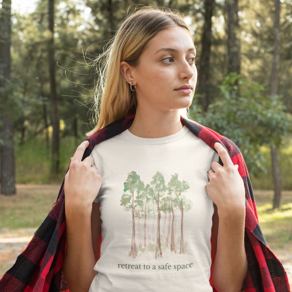 Woman wearing a t-shirt with a forest design and text, that says Retreat to a Safe Space, standing in a forest.